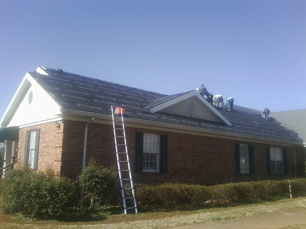 Crew tearing shingles off a brick ranch roof with a ladder set — Emergency Roof Repair work by our crew in Mc Donough