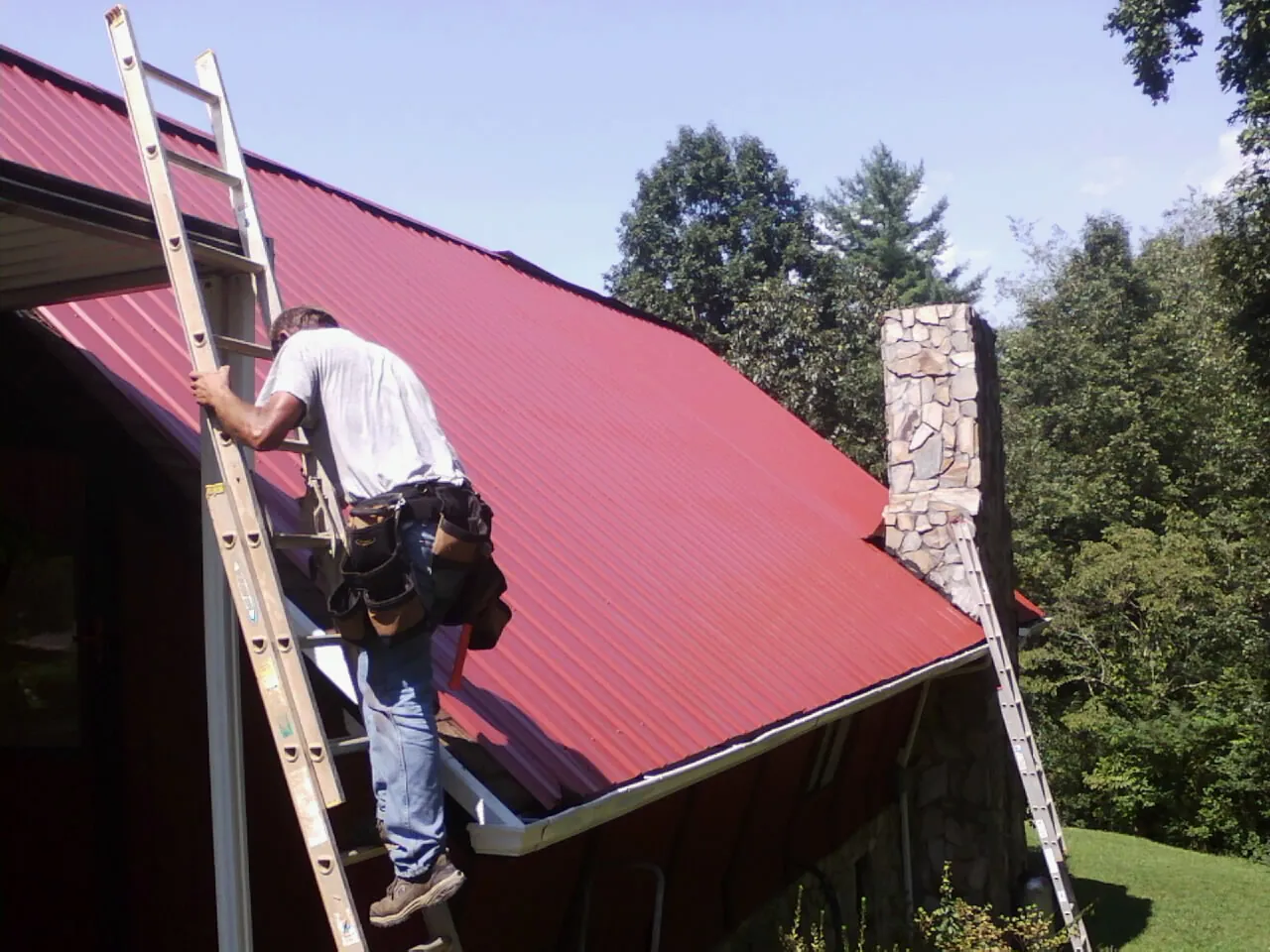 Roofer on a ladder beside a red metal roof and stone chimney — Soffit & Fascia Repair by Roofing Brothers in Mc Donough